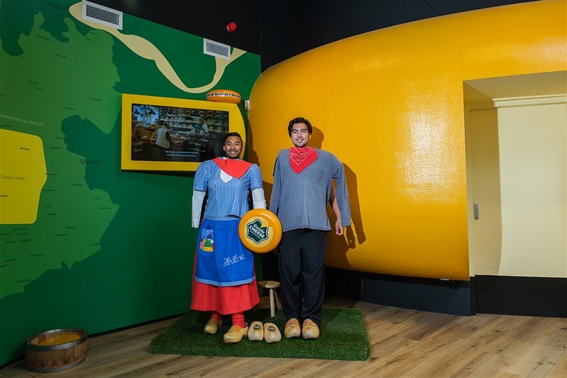Twee bezoekers poseren in traditionele Nederlandse kaasmarkt-kleding bij de Gouda Cheese Experience. Ze dragen klompen en staan voor een decor met een gigantische kaas en een interactieve kaart van Nederland.