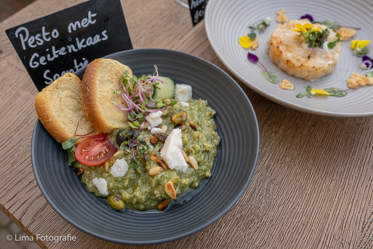 Bovenaanzicht van een culinair gerecht: groene pesto met brokjes witte geitenkaas in een donkere kom, gegarneerd met tomaat en micro-stengels, met een menukaartje "Pesto met Geitenkaas" ernaast.