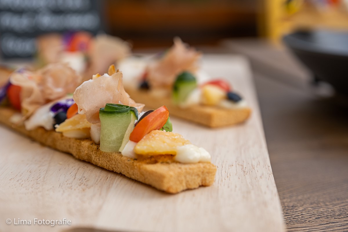 Een rij kleurrijke bruschetta's op een lichte houten serveerplank, gedecoreerd met ham, frisse groenten en eetbare bloemen, gefotografeerd met een geringe scherptediepte.